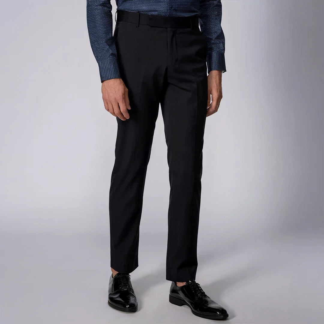 A man standing wearing black Kensington trousers with a mid-rise waist and sharp pleats, paired with a shirt and shoes.
