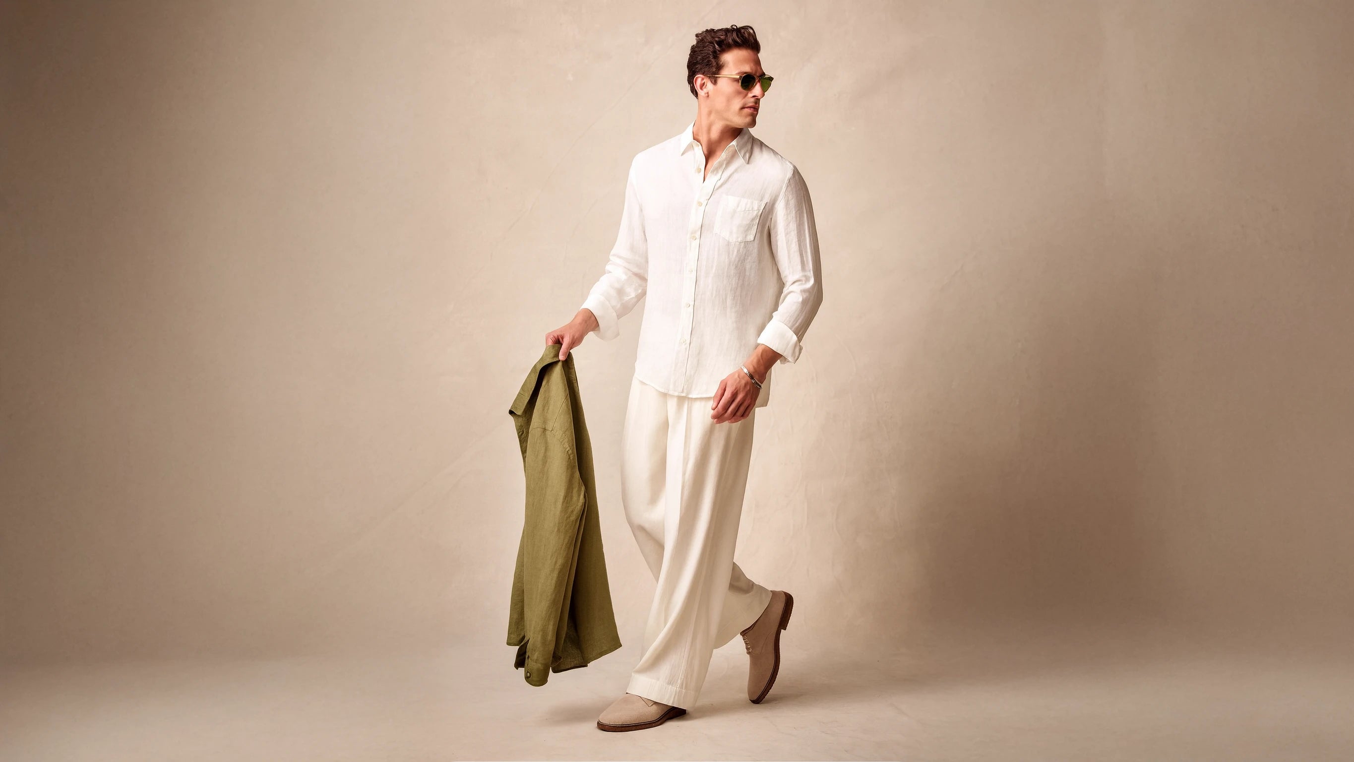 Man wearing a white linen shirt and white loose pants from House of Nirvana, holding a green linen shirt against a beige background.