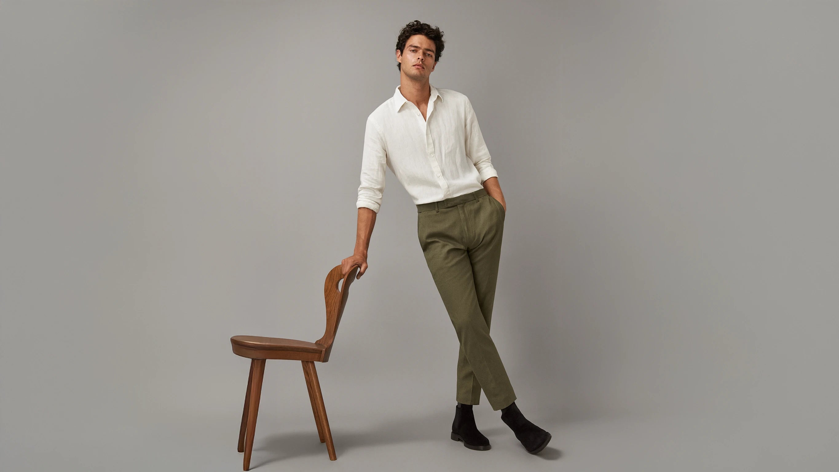 Man in a white linen shirt and green chinos standing next to a wooden chair on a grey background.