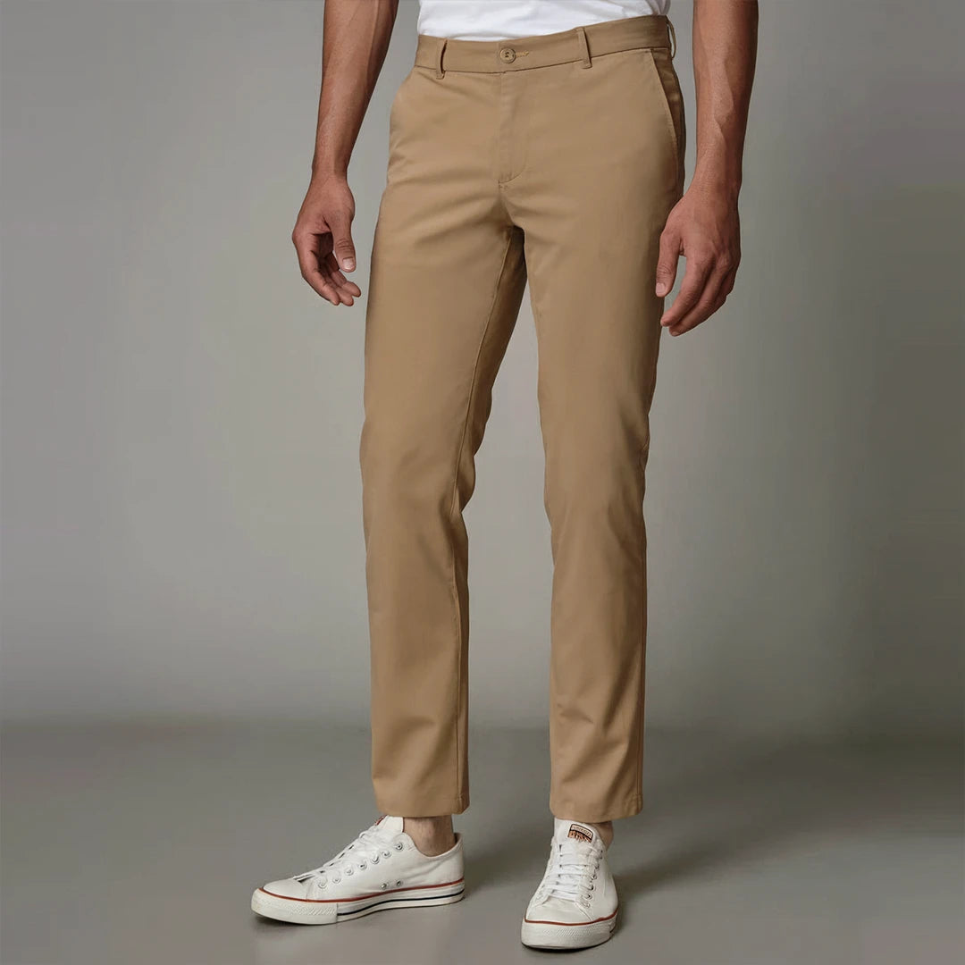A man standing wearing brown Kensington trousers with a mid-rise waist and sharp pleats, paired with a shirt and shoes.